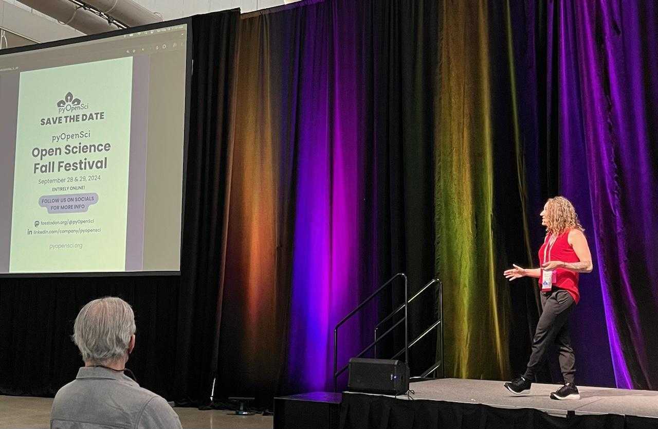 Image of a woman on a big stage wearing a red tank top talking. On the side is a slide that says pyOpenSci - Save the Date. Open science fall festival september 28-29 2024' style='width: 70%;